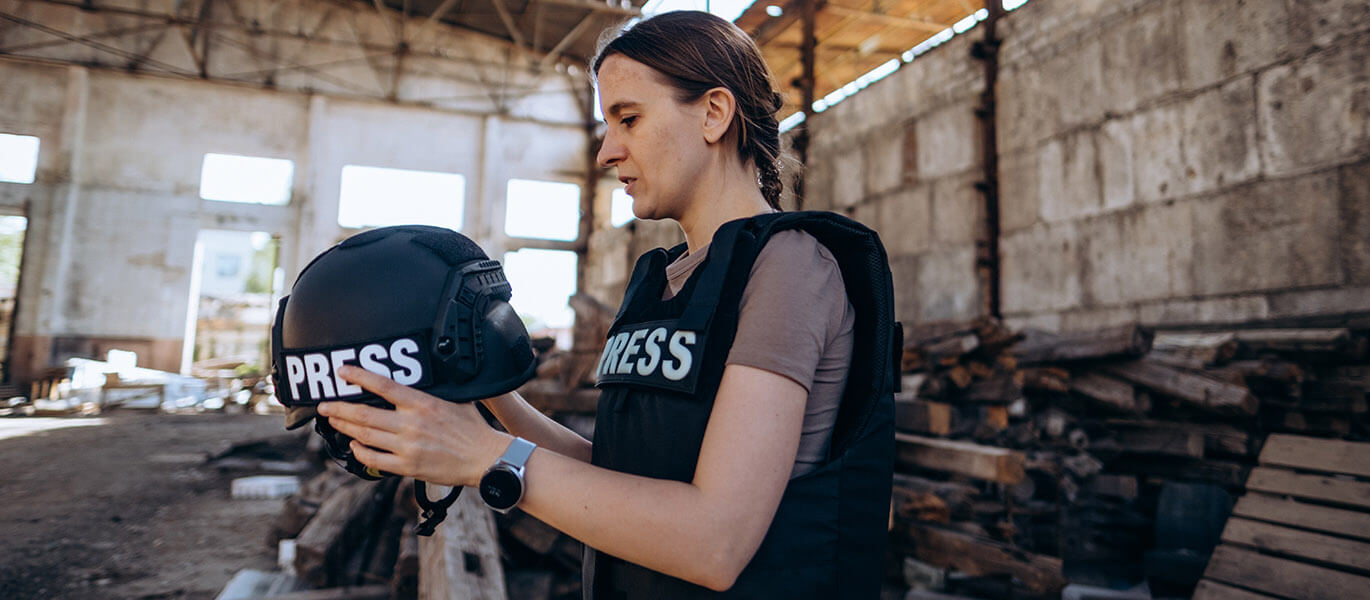 A Ukrainian woman journalist wearing a press vest and holding a press helmet. Behind her is a warehouse in disrepair.