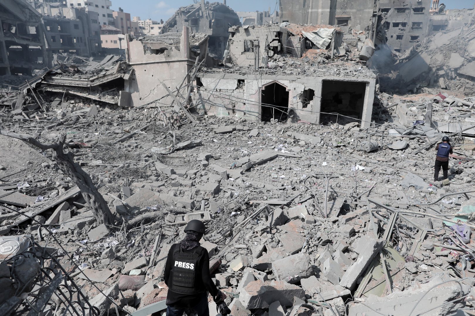 Two Palestinian journalists walk through the site of an airstrike. They are wearing press vests and are surrounded by grey ash and rubble.