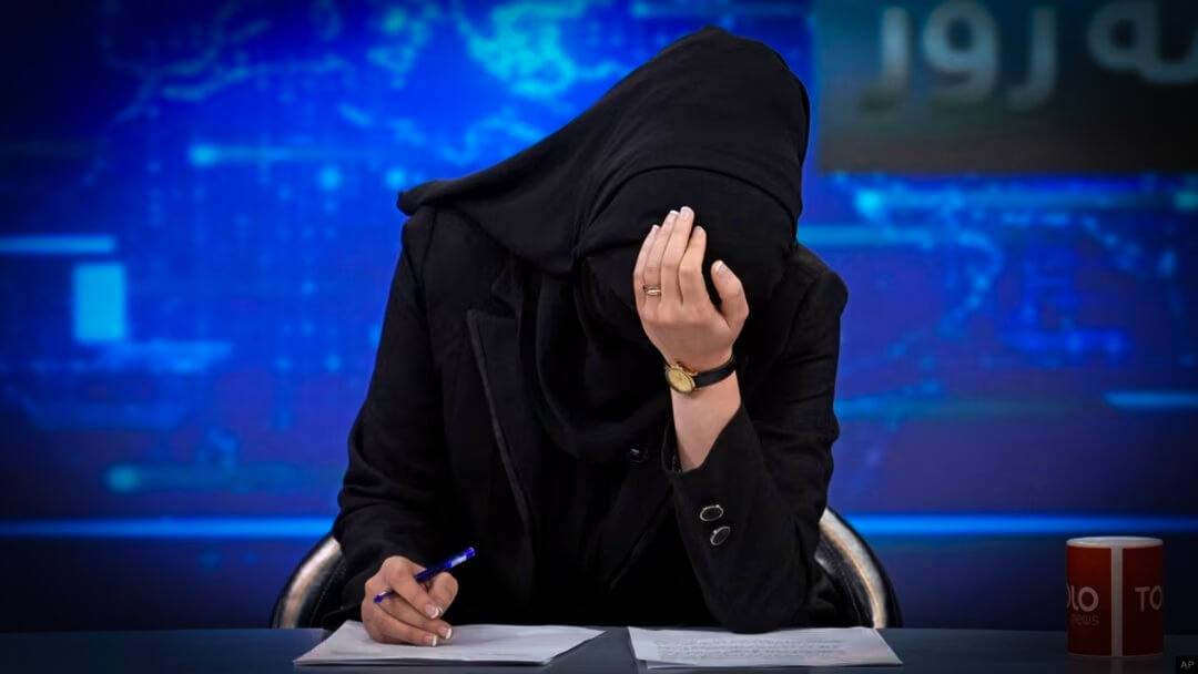 A woman working for Tolo News in Afghanistan sits at a news desk. She is fully veiled in black and holds her head in her hands - the only exposed skin visible.
