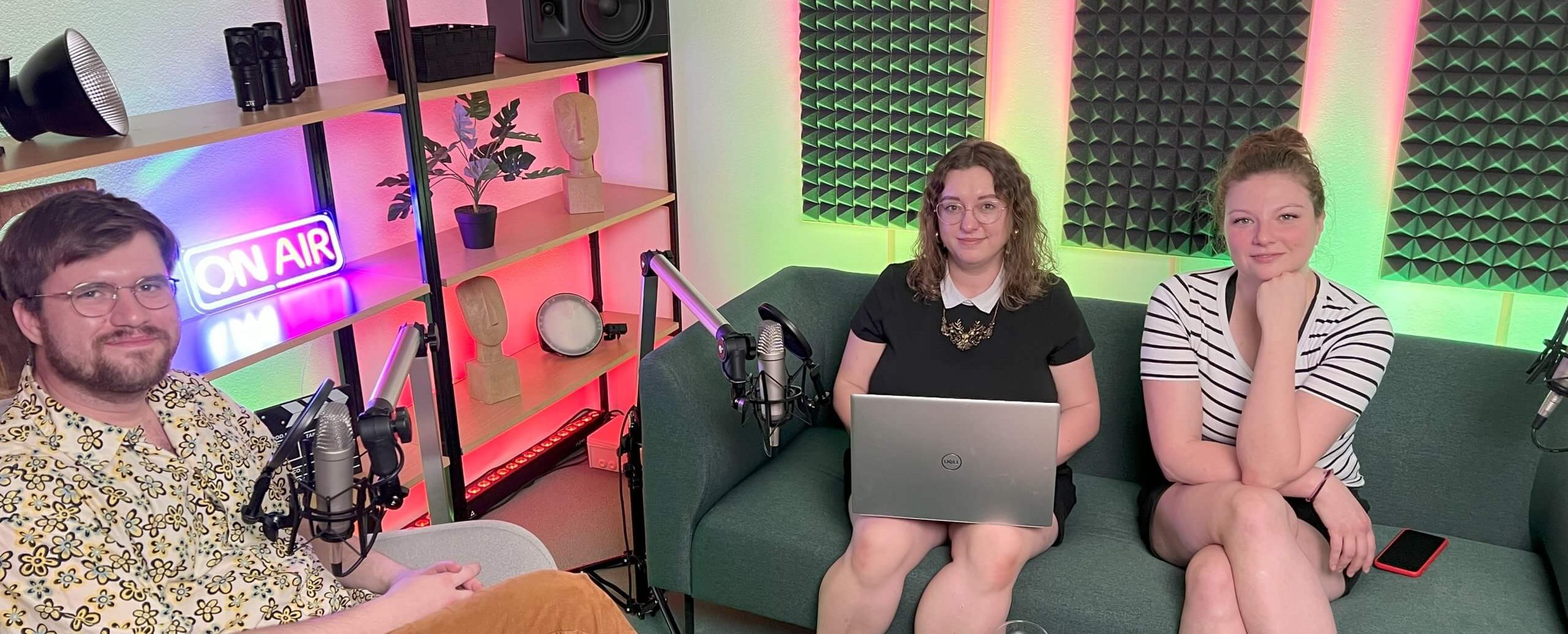 The team from Okraj, a man and two women, sit in a podcast studio with laptops and microphones.